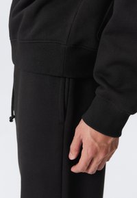 Nærbilde av en person som har på seg svarte joggebukser og en svart hettegenser med ribbestrikkede mansjetter, der de viser høyre hånd avslappet ved siden av kroppen.