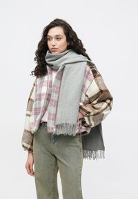 Sivý šál prehodený cez károvanú bomber bundu v ružových, hnedých a krémových tónoch, doplnenú o zelené džínsy. Šál má strapcové okraje.
