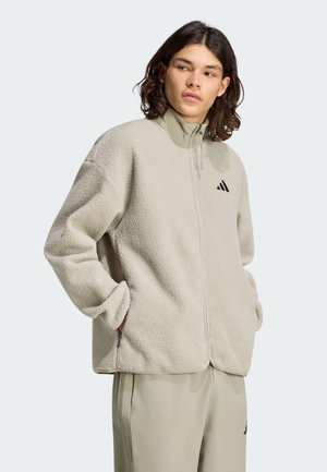 Persona con cabello hasta los hombros que lleva una chaqueta polar beige clara con cremallera y logo en el pecho, manos en los bolsillos, mirando de lado.