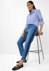 Donna con una camicia a righe blu e jeans con strisce laterali scure, seduta su uno sgabello alto, indossa scarpe piatte in rete nere e sorride.