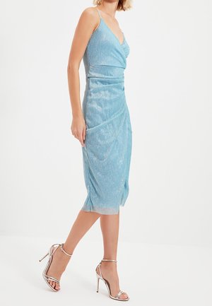 Frau trägt ein schimmerndes hellblaues ärmelloses Midikleid und metallisch silberne High-Heel-Sandalen, stehend vor weißem Hintergrund.