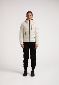 Piumino bianco con motivo a righe orizzontali, chiusura frontale con zip, due tasche laterali con zip e cappuccio. Indossato con pantaloni della tuta neri e sneakers.