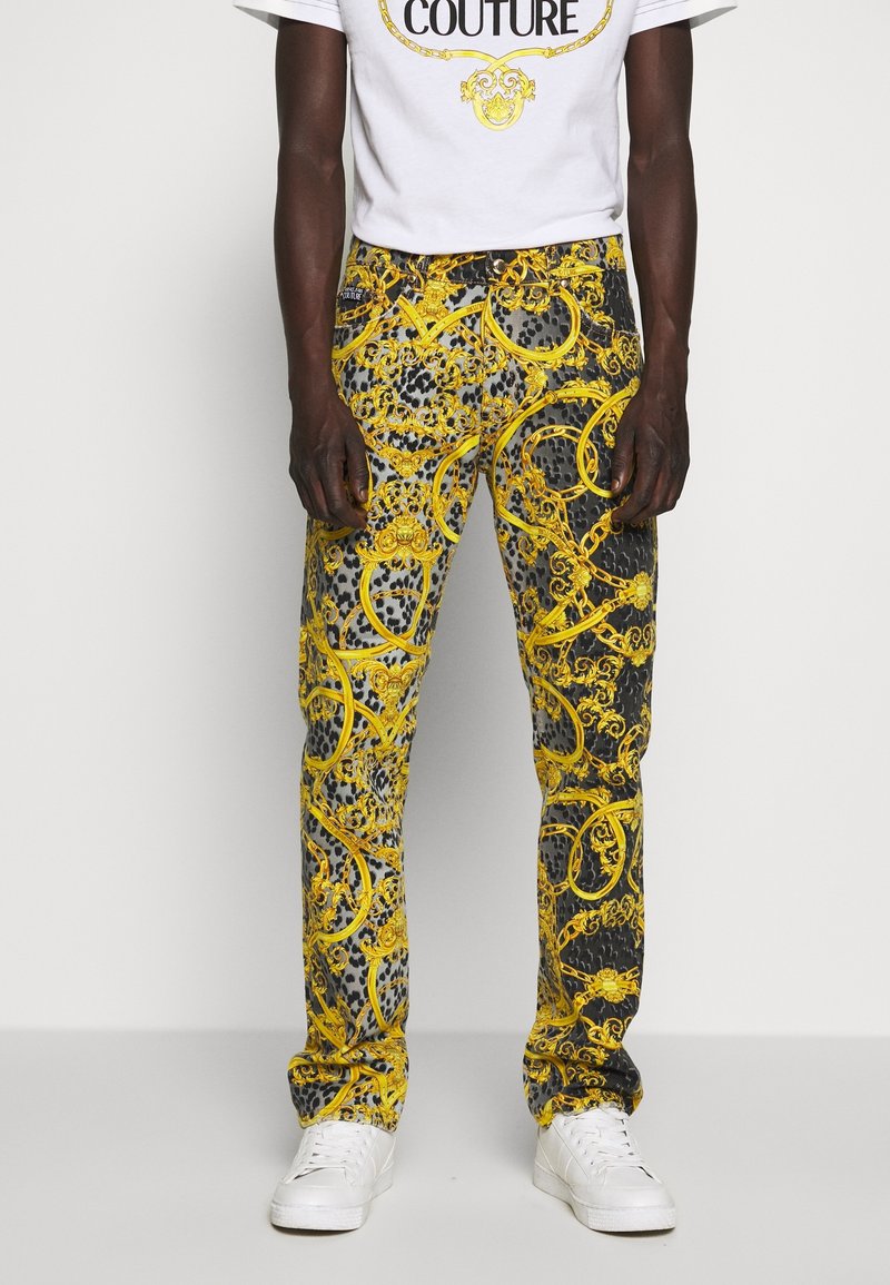 Gula och grå mönstrade byxor med ett tryck som visar kedjor och barockelement; slim fit med en mjuk textur, parat med vita sneakers.