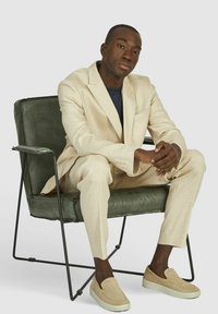 Beige linnen pak met een getailleerde pasvorm, witte instapschoenen, zittend op een groene leren en metalen stoel, neutrale achtergrond, ontspannen houding.