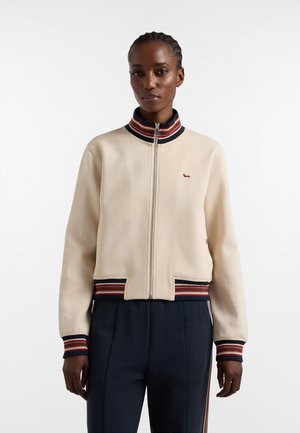 Donna che indossa una giacca beige con cerniera, colletto, polsini ed orlo a righe rosse e blu navy, abbinata a pantaloni blu navy, in piedi su sfondo bianco.