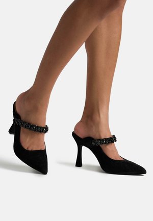 Mules neri in suede con tacco alto, caratterizzati da una punta affilata e una cinghia gioiello che attraversa il piede. La finitura testurizzata e la silhouette elegante esaltano il design.