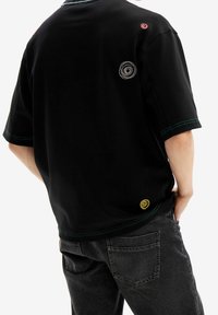 Persona vestida con una camiseta negra de gran tamaño con bordado de espiral colorido y jeans oscuros, de pie con una mano en el bolsillo.