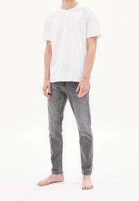Bílá bavlněná trička s krátkým rukávem v kombinaci se šedými slim fit džíny. Džíny mají vybledlý vzhled a kontrastní švy. Naboso.