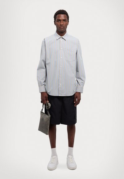 Camicia a maniche lunghe con righe verticali blu e bianche, con taschino sul petto, abbinata a pantaloni corti neri e borsa grigia. Sneakers bianche.