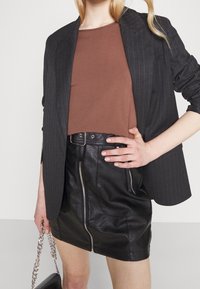 Jupe mini en cuir noir avec une fermeture éclair à l'avant, associée à un haut marron et un blazer sombre à rayures. Tenue avec un sac à chaînes.