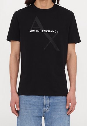 Person trägt ein schwarzes Armani Exchange T-Shirt mit großem weißen Text "AX" und "ARMANI EXCHANGE" auf der Brust, kombiniert mit blauen Jeans.