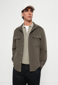 Mann trägt eine beigen Mütze, ein olivfarbenes, geknöpftes Hemd über einem hellgrauen V-Ausschnitt-Pullover und schwarze Hosen, steht mit den Händen in den Taschen.
