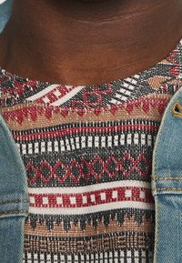 Gebreide shirt met patroon in tinten van rood, zwart, wit en beige; heeft geometrische ontwerpen. Mouw van een blauwe spijkerjack staat aan de linkerkant.