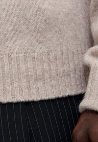 Jersey de punto beige con dobladillo acanalado y textura suave, combinado con pantalones negros con rayas para crear contraste.