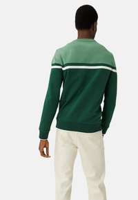 Sudadera verde y verde azulado con un diseño a rayas en la parte trasera; puños y dobladillo acanalados; combinada con pantalones de color claro.