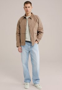 Bruine microvezel jas met drukknoopsluitingen, geribde kraag, voorzakken, gedragen over een lichtgroen shirt, gecombineerd met lichtblauwe spijkerbroek en witte sneakers.