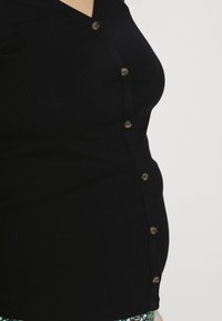 Närbild av en person som bär en svart ribbad knäppt cardigan, som visar sidan av överkroppen från axel till höft mot en vit bakgrund.