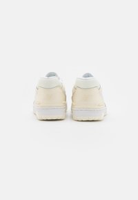 Un paio di sneakers beige e bianche New Balance visto da dietro su una superficie bianca e liscia.