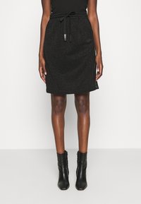 Mujer vestida con una falda negra de tiro a la rodilla con un brillo sutil, combinada con botas de tobillo negras de punta, de pie frente a un fondo neutro.