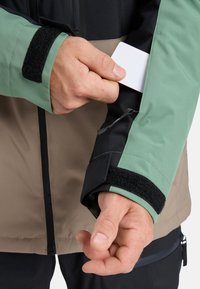 Waterproof jacket with green and black sleeves. Features Velcro cuff, zipper, and pocket for card storage. Design includes beige torso.