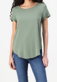 Sagegrøn kortærmet top i et let materiale. Har en rund halsudskæring og en buet kant, parret med blå jeans.