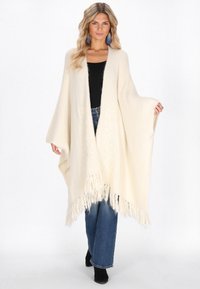 Poncho crem cu margine franjurată, textură moale, design deschis în față, asortat cu blugi închiși și cizme negre până la gleznă.