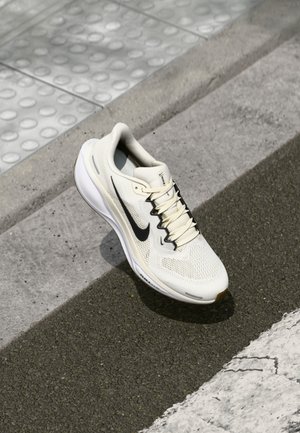 Baskets de sport blanches avec logo Swoosh noir placées sur un pavé extérieur texturé et un bord de chaussée.