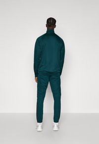 Zeleni trenirki z visokim ovratnikom in dolgimi rokavi, z ozkim krojem. Hlače imajo stranske črte in eleganten dizajn, v kombinaciji z belimi supergami.