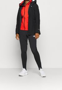 Person, der eine schwarze Jacke, ein rotes Zip-Up-Oberteil, schwarze Leggings mit Logo, schwarze Socken und weiße Turnschuhe trägt, steht vor einem schlichten weißen Hintergrund.