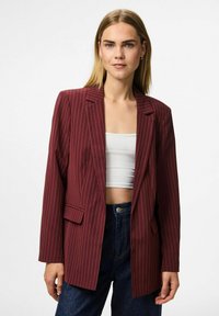 Young woman wearing a burgundy pinstripe blazer over a white crop top and dark blue jeans, standing against a plain white background.