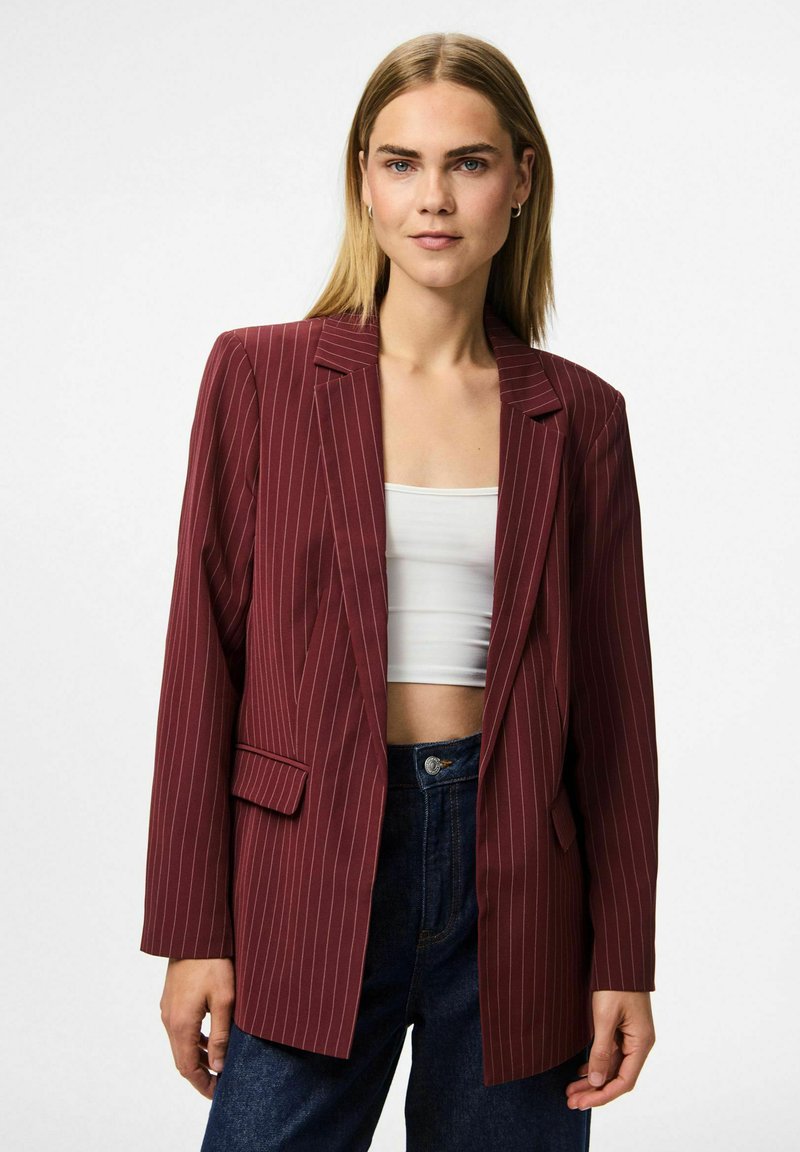 Young woman wearing a burgundy pinstripe blazer over a white crop top and dark blue jeans, standing against a plain white background.
