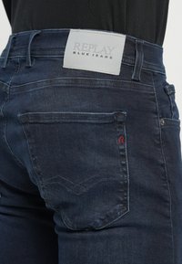 Mørkeblå denim jeans med en grå label med teksten "REPLAY BLUE JEANS", rødt syning detalje og et særligt baglommelys design.
