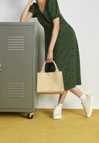 Femme en robe fleurie verte et baskets blanches s'appuyant contre un casier en métal gris, tenant un sac à main beige contre un mur blanc et un sol jaune.