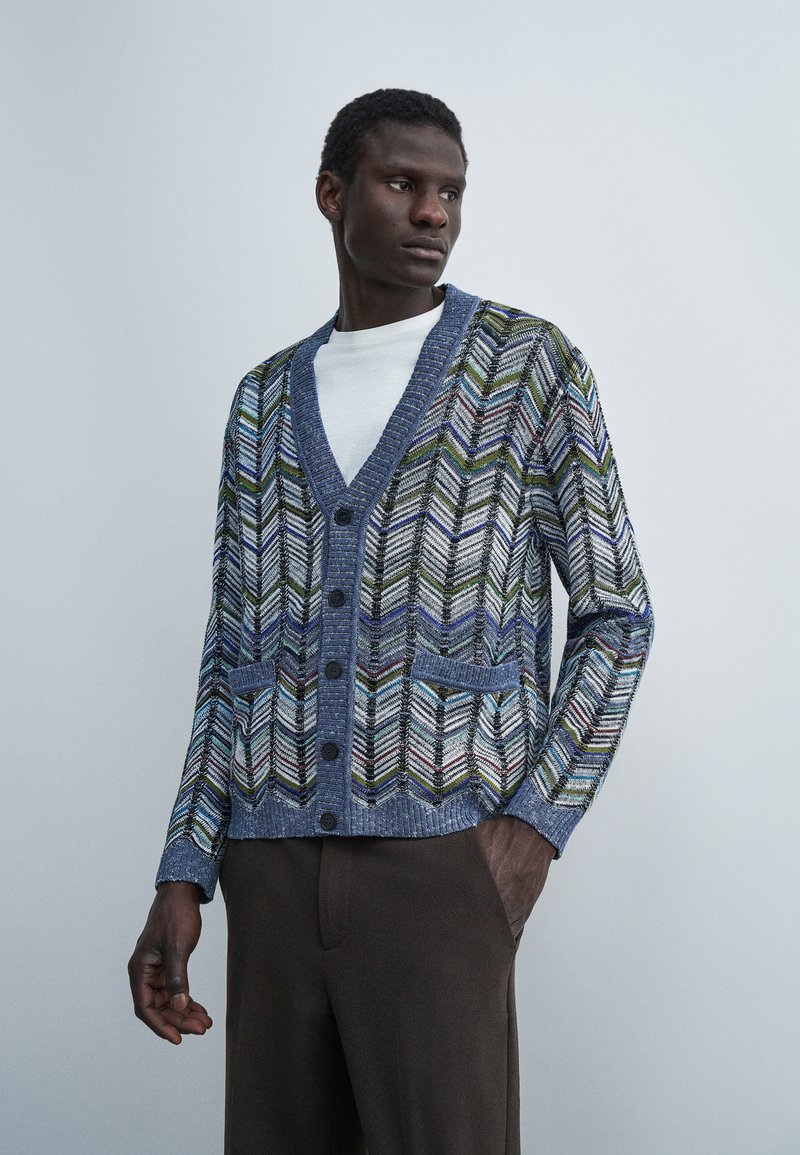 Άνδρας που φοράει μπλε πλεκτή ζακέτα με μοτίβο και κουμπιά πάνω από λευκό πουκάμισο και σκούρο παντελόνι, κοιτάζοντας στο πλάι με το ένα χέρι στην τσέπη.