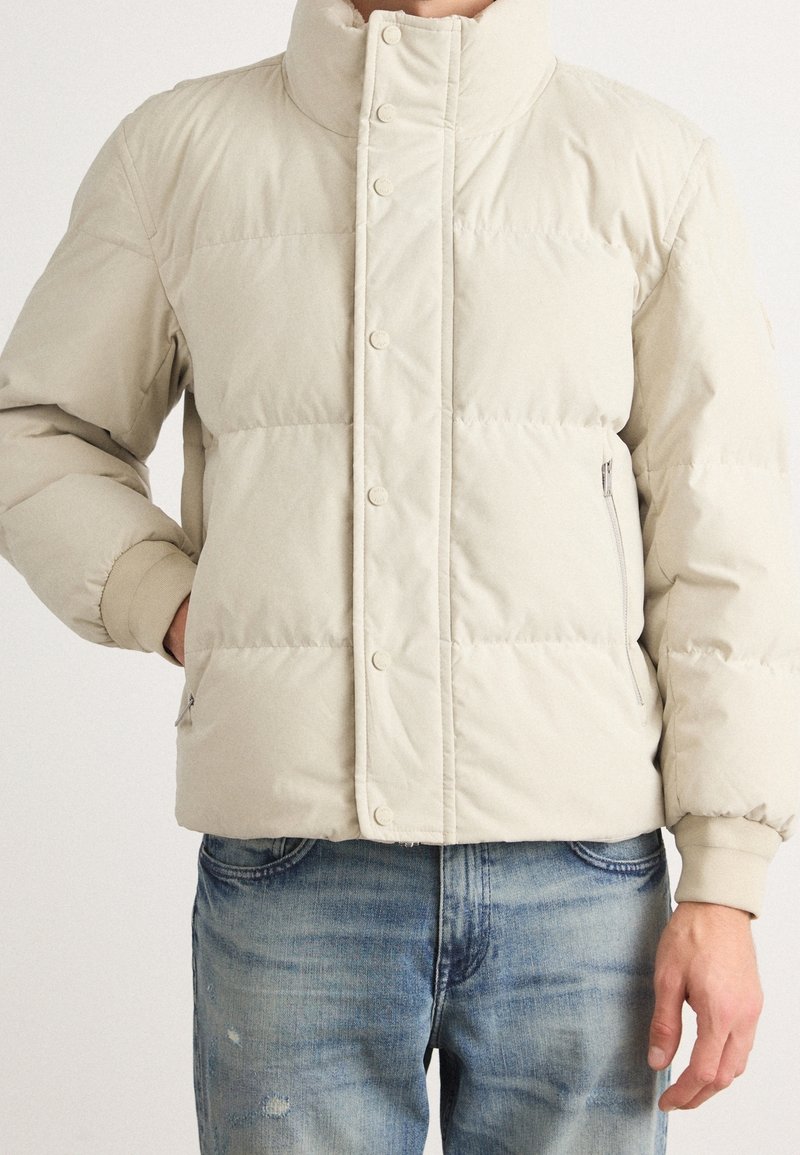 Doudoune beige avec boutons-pression, poches zippées, poignets côtelés, portée avec un jean bleu clair délavé.