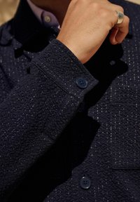 La giacca textured blu navy presenta un motivo intrecciato, due bottoni blu sui polsini e una vista ravvicinata del colletto con tessuto a contrasto.