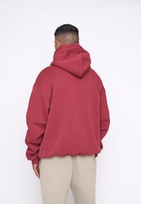 Maroon hoodie with a large drawstring hood, loose fit, and ribbed hem. The fabric appears soft and textured, worn over beige pants.