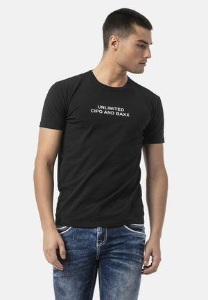 Mann trägt ein schwarzes T-Shirt mit kurzen Ärmeln und dem Text "UNLIMITED CIPO AND BAXX", kombiniert mit blauen Jeans, schaut nach rechts.
