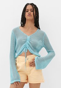 Himmelblauer, kurz geschnittener Strickpulli mit langen, weiten Ärmeln und Kordelzugdetails an der Taille, kombiniert mit pfirsichfarbenen Shorts.