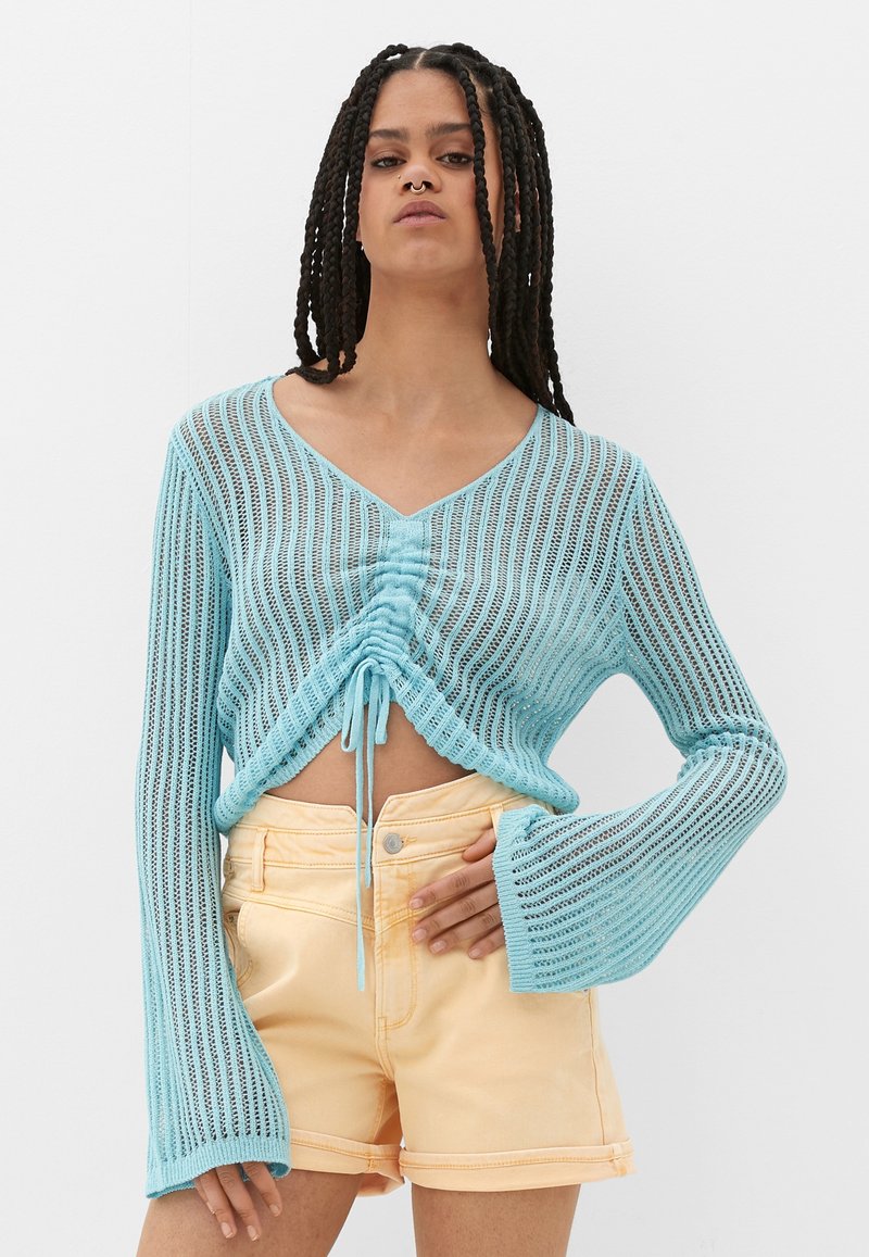 Himmelblauer, kurz geschnittener Strickpulli mit langen, weiten Ärmeln und Kordelzugdetails an der Taille, kombiniert mit pfirsichfarbenen Shorts.