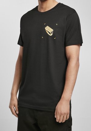 Mann trägt schwarzes T-Shirt mit goldfarbenem Ringgrafik und dem Wort "PRAY" auf der oberen linken Brust bedruckt.