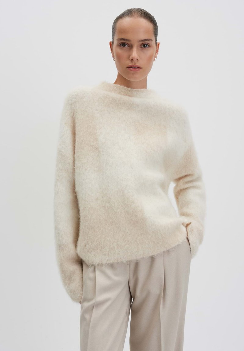Cream-colored, fuzzy sweater with a high neckline and long sleeves, paired with light beige, pleated trousers. Soft, textured fabric.