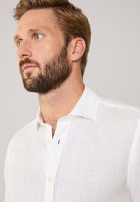 Witte linnen blouse met een gespreide kraag, voorzien van een knoopsluiting en subtiele textuur, gedragen door een man met een baard en lichtbruin haar.