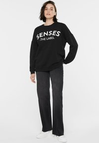 Schwarzer Sweatshirt mit weißem Text "SENSES .THE LABEL," kombiniert mit dunklen Jeans und weißen Sneakers. Lockere Passform und weicher Stoff.