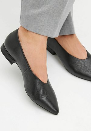 Chaussures en cuir noires à bout pointu et talons bas portées avec un pantalon gris clair, pieds nus, sur fond blanc.