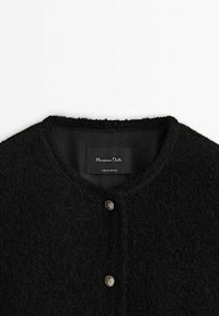 Svart ullkofta med rund halsringning, subtil textur och två knappar i silverfärg framtill. Etiketter synliga inuti.