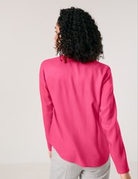 Fuchsia blouse with long sleeves, smooth texture, rounded hemline, and a slightly loose fit, paired with light gray trousers.