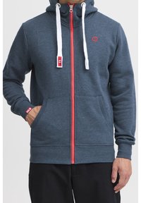 Uomo che indossa una felpa con zip blu navy con zip rossa, lacci bianchi e con una mano nella tasca frontale, mostrato dalla spalla ai fianchi.