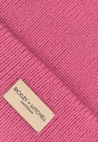 Felroze gebreide stof met een textuurpatroon. Een beige label met de tekst "BICKLEY + MITCHELL AMSTERDAM," geborduurd in het zwart.