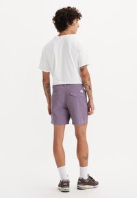 Shorts mov cu un amestec de bumbac, de culoare purpurie, cu un buzunar pe spate și detalii subtile de cusătură, asortați cu un tricou alb și încălțăminte sport.